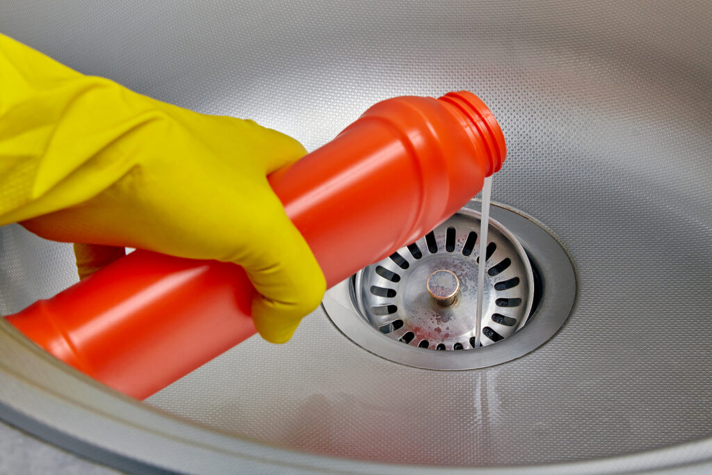 Gloved hand pouring chemical pipe cleaner down a stainless steel kitchen sink drain