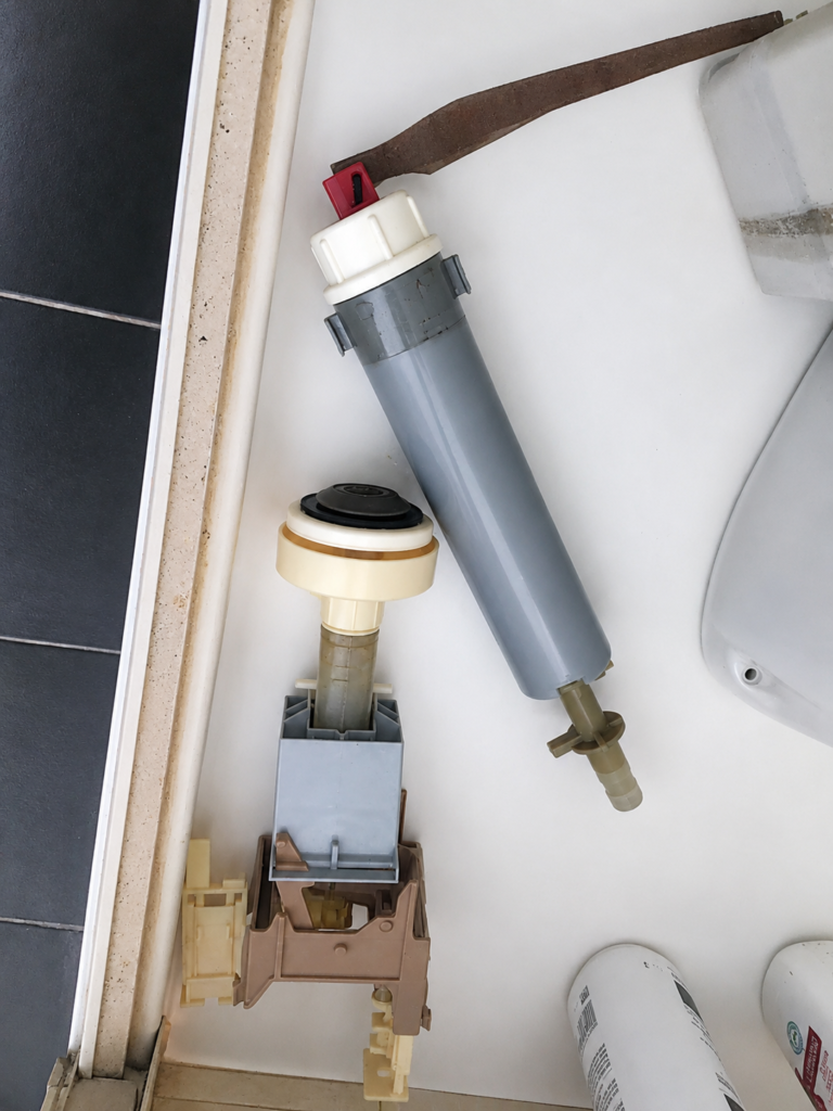 Old inlet and outlet valves removed from a toilet cistern, laid on the bathroom floor beside the pan